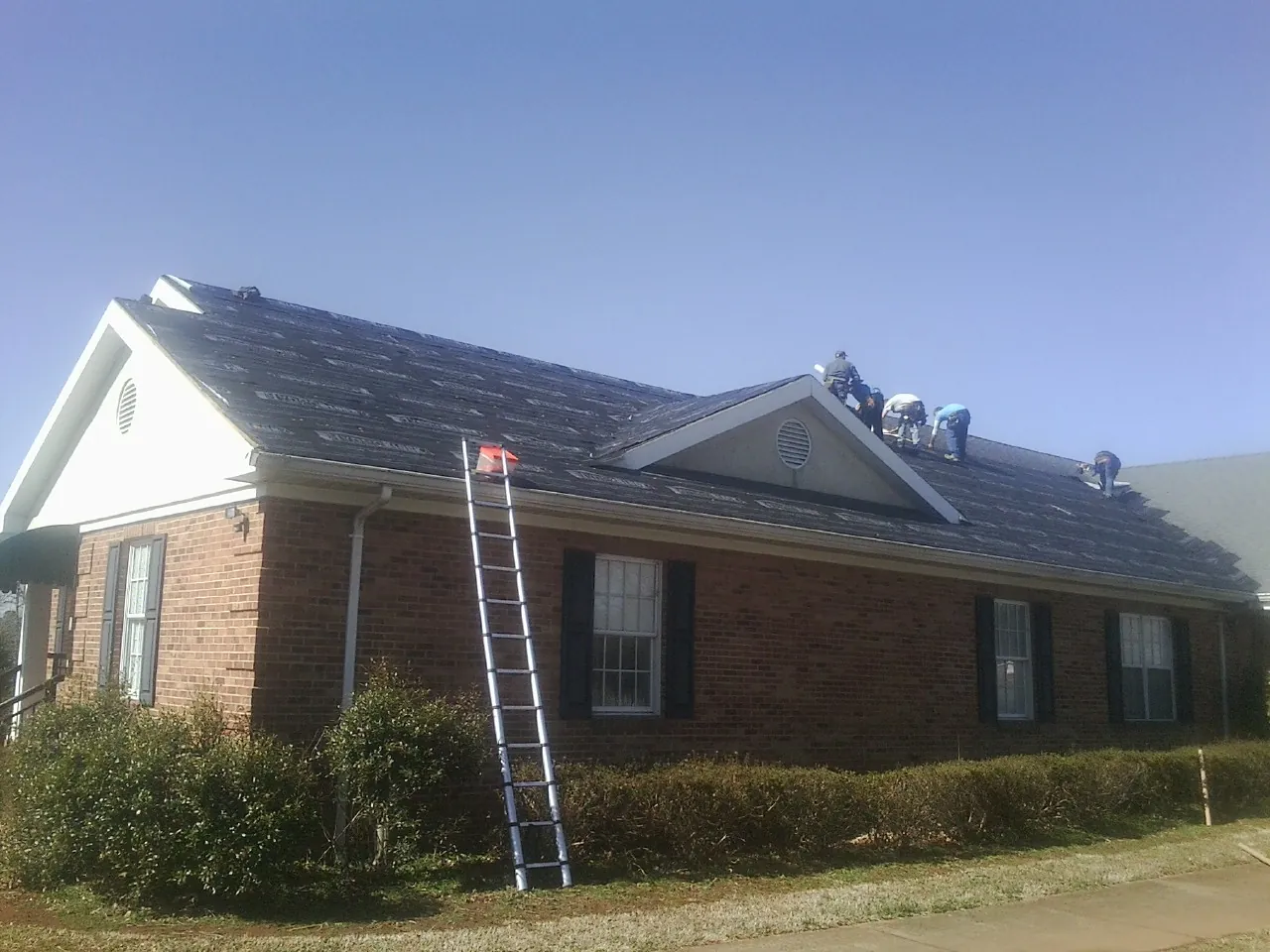 Crew tearing shingles off a brick ranch roof with a ladder set — Metal Roof Repair work by our crew in Neola