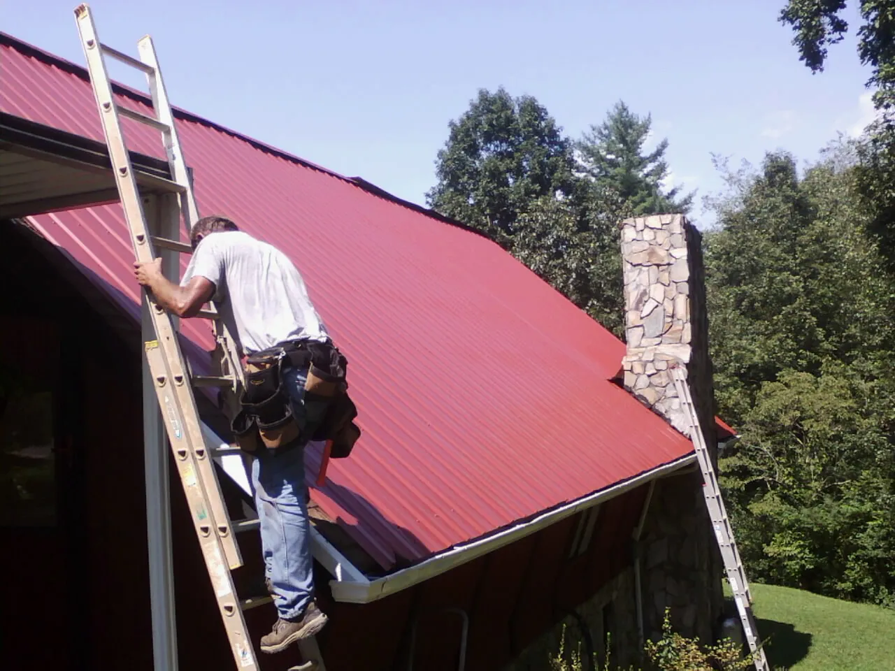 Roofer on a ladder beside a red metal roof and stone chimney — Soffit & Fascia Repair by Roofing Brothers in Neola