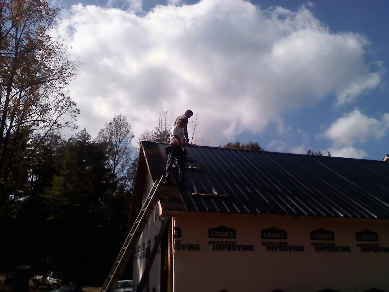 Roofer fastening dark metal panels high against a cloudy sky, from a Roof Inspection job in Neola